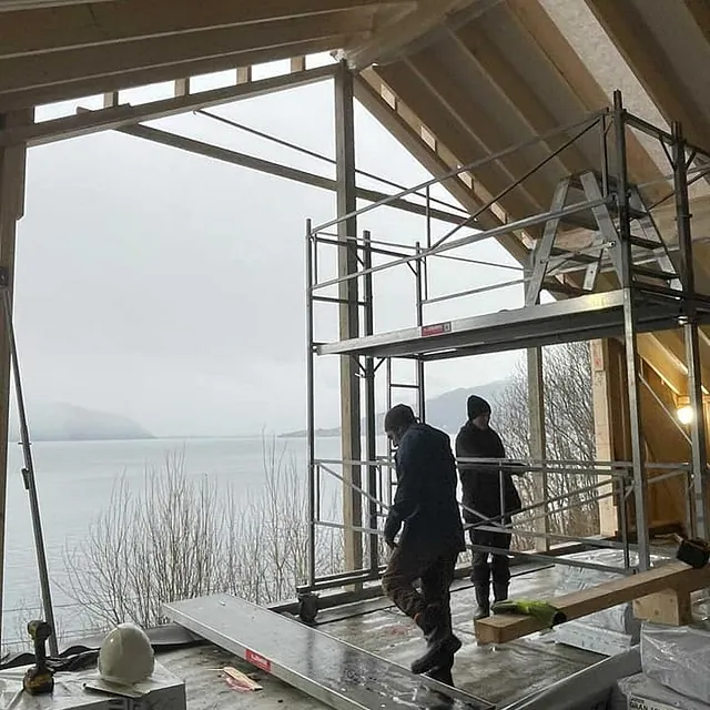 To arbeidere med stillas monterer husramme med sjøutsikt i bakgrunnen.