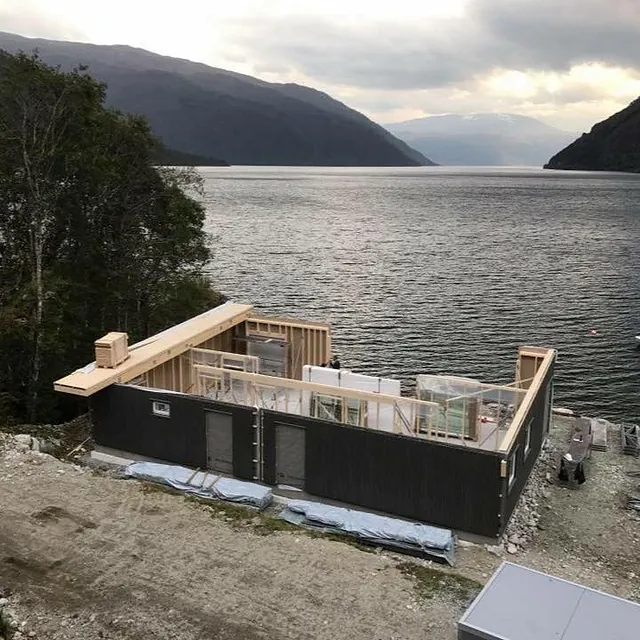 Hus under oppføring ved fjord, yttervegger delvis reist med åpen takramme.